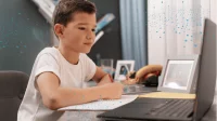 Ein Junge sitzt am Schreibtisch und schreibt eine mit KI generierte Hausaufgabe von seinem Laptop ab