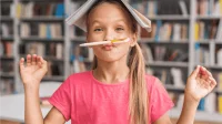 Foto von einem lustigen Mädchen, dass sich im Unterricht ein Buch auf den Kopf gesetzt und einen Stift unter die Nase geklemmt hat