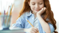 Grundschülerin sitzt an einem Schreibtisch und schreibt mit einem Stift in ein Heft