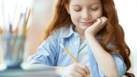 Grundschülerin sitzt an einem Schreibtisch und schreibt mit einem Stift in ein Heft