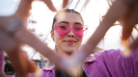 Eine junge Frau schaut mit rosa Sonnenbrille in die Kamera und stellt ein Herz mit ihren Fingern dar