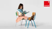 Kind sitzt auf einem Stuhl und liest während es die Beine auf einem anderen Stuhl abgelegt hat