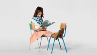 Kind sitzt auf einem Stuhl und liest während es die Beine auf einem anderen Stuhl abgelegt hat