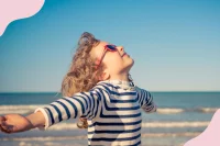 Ein Kind mit Sonnenbrille streckt am Strand die Arme weit aus