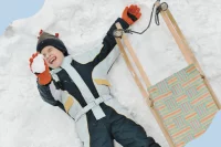 Ein Kind liegt neben seinem Schlitten im Schnee und hält einen Schneeball nach oben