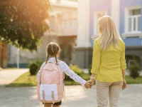 Mutter und Kind mit Schulranzen stehen Hand in Hand vor einer Schule