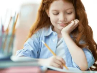 Grundschülerin sitzt an einem Schreibtisch und schreibt mit einem Stift in ein Heft