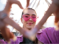 Eine junge Frau schaut mit rosa Sonnenbrille in die Kamera und stellt ein Herz mit ihren Fingern dar